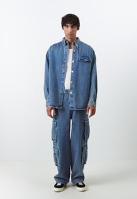 Casaco de ganga com botões, bolso frontal e um corte descontraído, combinado com jeans cargo de pernas largas que apresentam múltiplos bolsos laterais em azul claro.