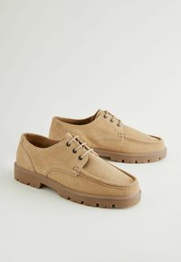 Zapatos de ante beige con punta redondeada, diseño de dos ojetes y suela de goma con tracción. Presenta detalles cosidos a lo largo de la parte superior.