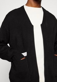 Cárdigan negro de corte relajado, con puños acanalados, abertura frontal y un bolsillo con detalle de etiqueta. Se lleva sobre una camisa blanca.