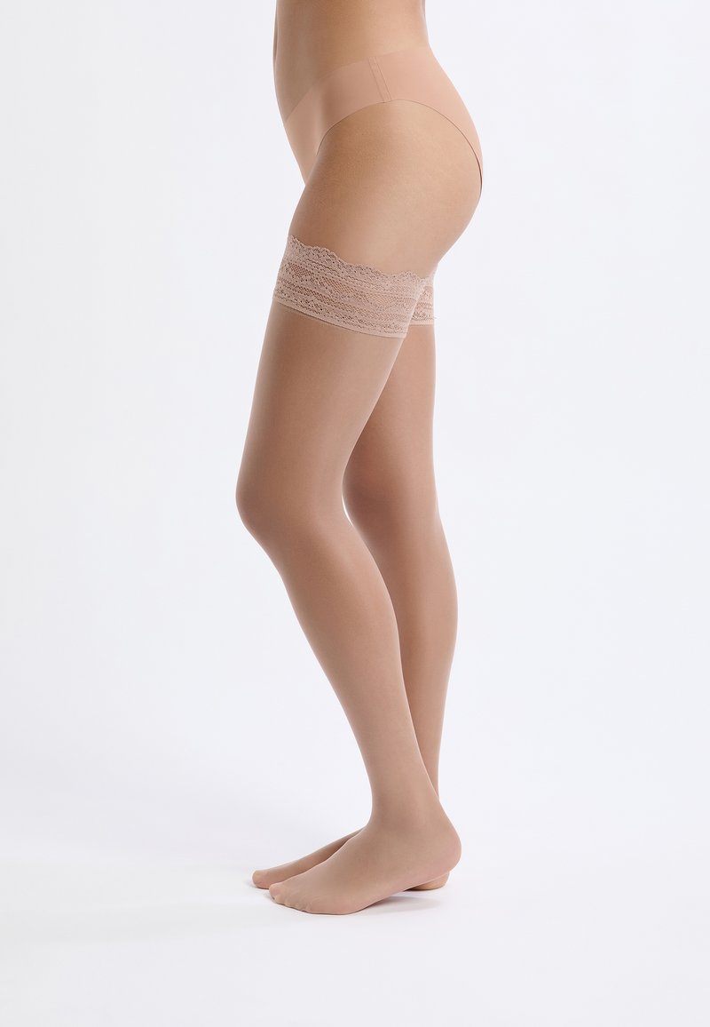 Przezroczyste beżowe pończochy do ud z kwiecistymi koronkami na górze, o gładkiej strukturze i dopasowanym kroju, spoczywające nad bosymi stopami.
