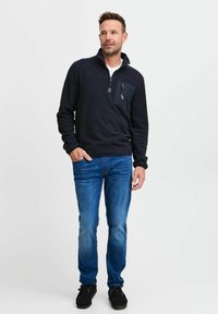 Navy Pullover mit einem Viertel-Reißverschluss-Kragen, ausgestattet mit einer schwarzen Brusttasche, über einem weißen Hemd getragen, kombiniert mit blauen Jeans und schwarzen Schuhen.