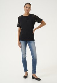 Melnas blūzes ar mežģīņu īsajām piedurknēm, savilktā kakla izgriezumā, kombinēta ar zilajiem pieguļošajiem džinsiem un melnajām kleitu kurpēm, stāvot pret baltu fona.