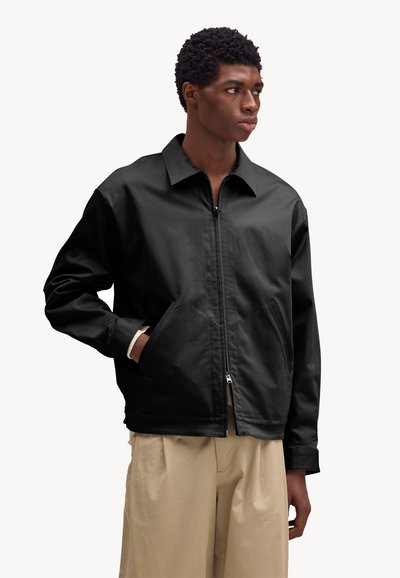 Giovane uomo che indossa una giacca nera con zip, con le mani nelle tasche e pantaloni beige a gamba larga, che guarda di lato su uno sfondo semplice.