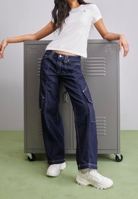 Hvid t-shirt med logo; mørkeblå brede cargo jeans med hvidt syning; parret med hvide sneakers; sat mod et gråt skab.