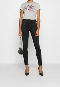 Camiseta gris con estampado floral, vaqueros negros de cintura alta con tachuelas decorativas, tacones negros en punta y un pequeño bolso cruzado negro.