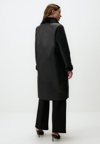 Long manteau noir avec un col et des poignets en fausse fourrure. Texture lisse, coupe droite et poches latérales. Accompagné d'un pantalon noir à jambes larges.