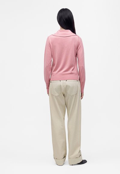 Femme aux longs cheveux noirs portant un haut rose à manches longues et un pantalon beige à jambes larges, debout, de dos, sur un fond blanc uni.