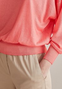 Osoba w koralowym swetrze z długim rękawem i ściągaczami oraz beżowych spodniach, z jedną ręką włożoną do kieszeni.