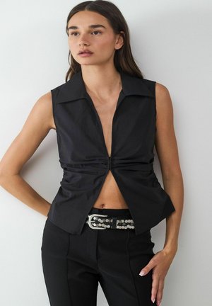 Schwarzes ärmelloses Shirt mit tiefem V-Ausschnitt, gerafftem Vorderteil und spitzem Kragen, kombiniert mit maßgeschneiderten schwarzen Hosen und einem Nieten-Gürtel.