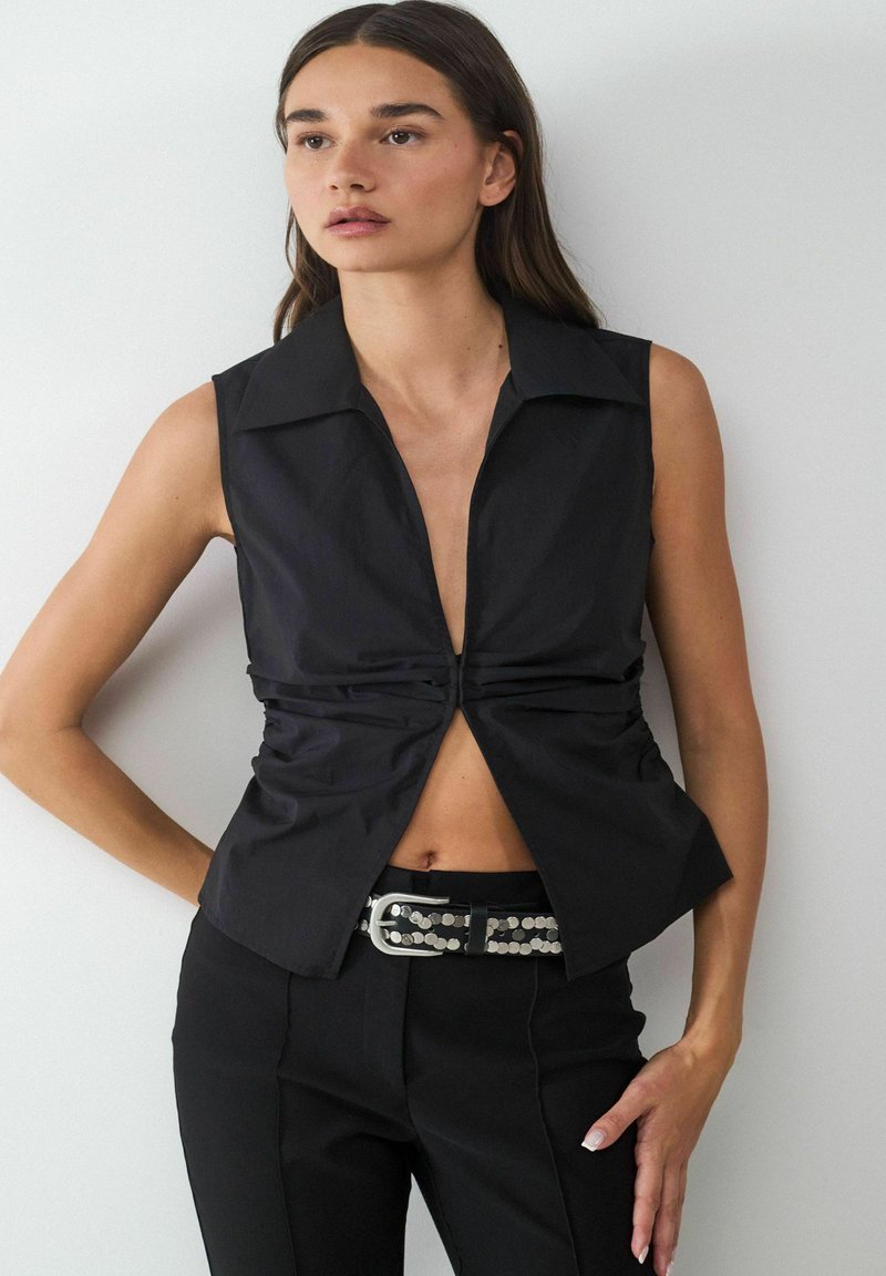 Schwarzes ärmelloses Shirt mit tiefem V-Ausschnitt, gerafftem Vorderteil und spitzem Kragen, kombiniert mit maßgeschneiderten schwarzen Hosen und einem Nieten-Gürtel.