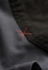 Tissu texturé noir et gris avec un détail brodé en rouge en forme de croix au niveau de la couture, mettant en valeur le contraste et le style de couture.