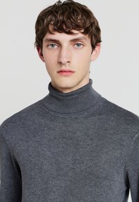 Grauer Rollkragenpullover aus weichem Stoff, mit einem hohen Kragen und gerippten Abschlüssen am Kragen, der eine taillierte Silhouette bietet.