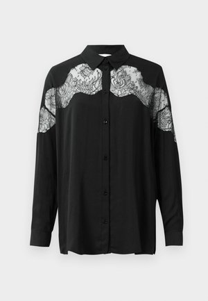 Chemise noire à manches longues. Présente un superposition en dentelle sur les épaules, ajoutant texture et contraste de motifs au tissu.