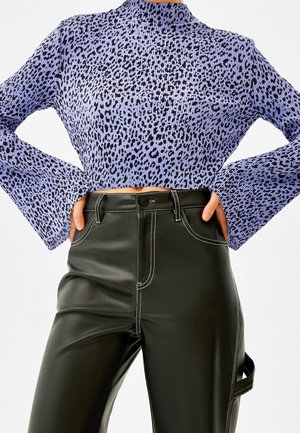 Frau trägt ein lila Oberteil mit Leopardenmuster, langen Ärmeln mit ausgestellten Manschetten, und hoch taillierte schwarze Kunstlederhose mit weißen Nähten.