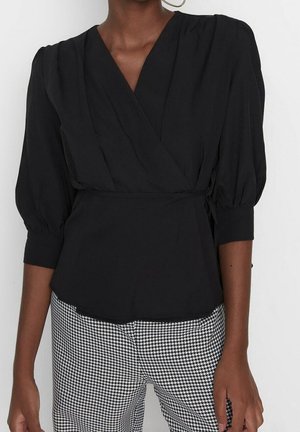 Vrouw draagt een zwarte wikkelblouse met driekwart ballonmouwen en zwart-witte pied-de-poule broek, staand tegen een witte achtergrond.