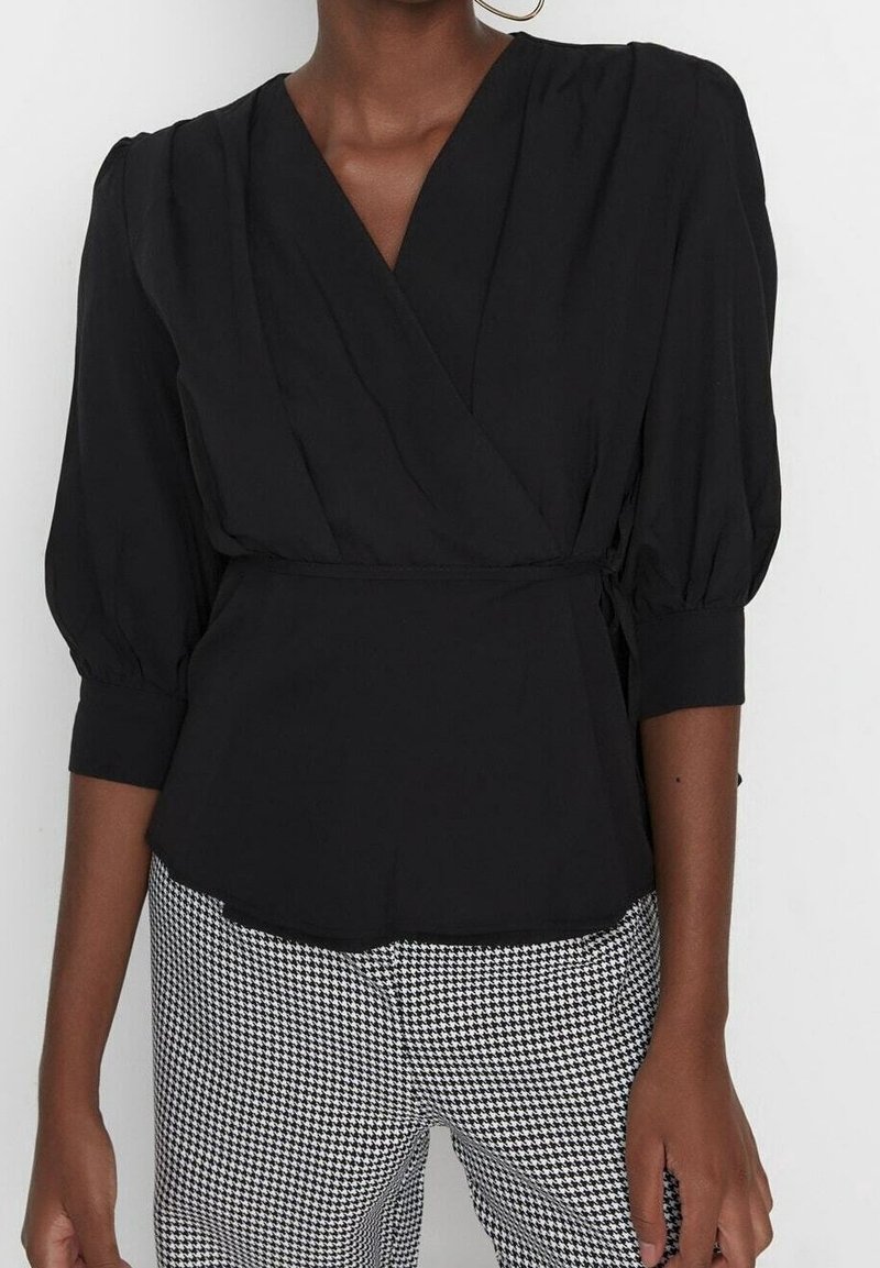 Femme portant un chemisier noir à nouer avec des manches trois-quarts bouffantes et un pantalon à motif pied-de-poule noir et blanc, debout devant un fond blanc.