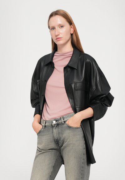 Camisa de cuero negro de gran tamaño con bolsillos frontales, superpuesta sobre un top de cuello alto en un suave tono rosa. La modelo lleva puestos unos jeans gris de cintura alta.