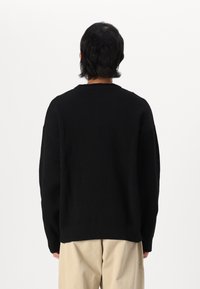 Suéter negro de punto acanalado con cuello redondo, mangas largas y un corte relajado. Llevado con pantalones de color claro, visto desde atrás.
