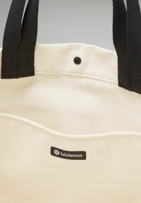 Canvas boodschappentas in crèmewit met een zwart logo, voorzien van zwarte stoffen handvatten en een voorzak met een drukknoopsluiting.