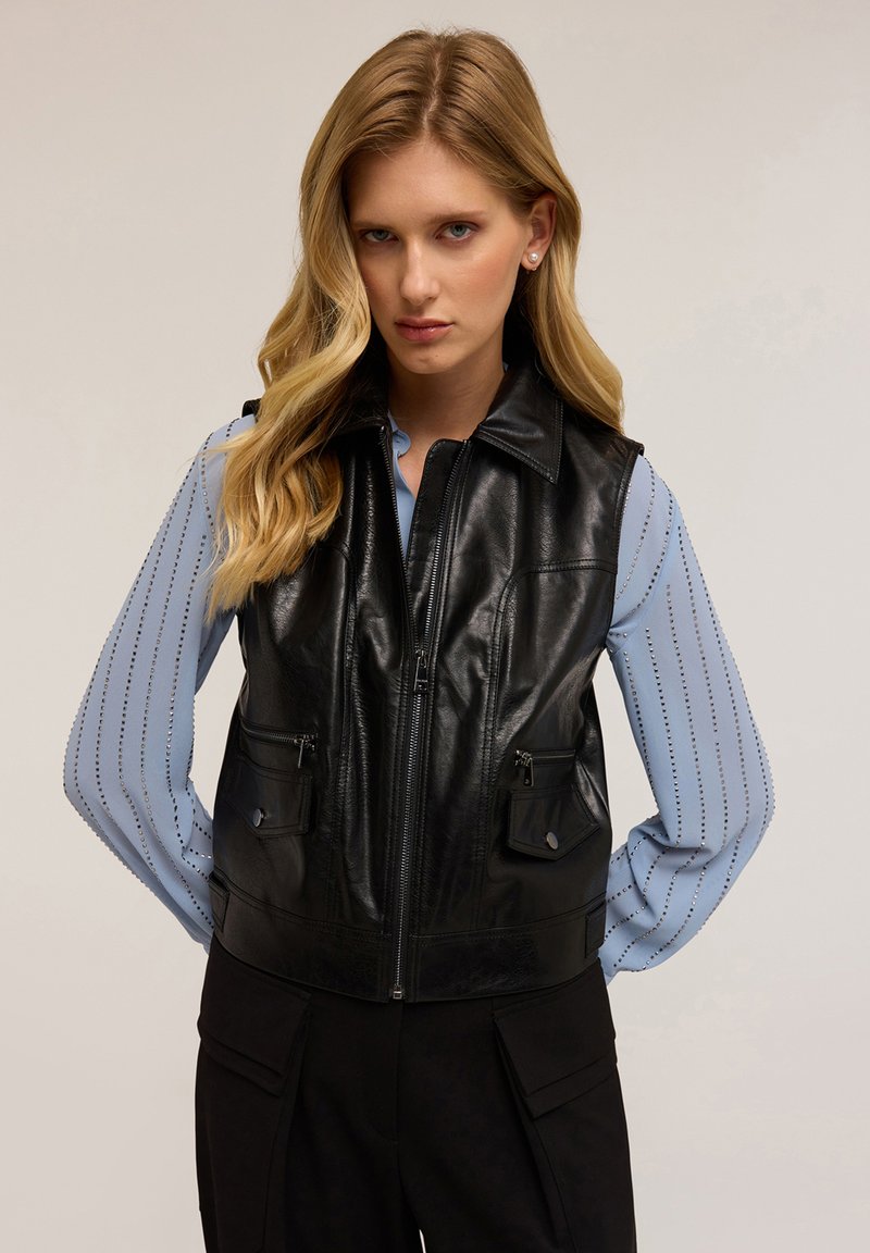 Czarna skórzana kamizelka bez rękawów z zamkiem z przodu, dwoma kieszeniami na klapie i spiczastym kołnierzem; noszona na jasnoniebieskiej koszuli w paski.