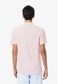 Jasnoróżowa koszulka polo, krótki rękaw, z kołnierzykiem, gładka tekstura i prosty dół, noszona przez osobę odwróconą tyłem.