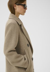 Abrigo de lana beige con cuello de muesca, diseño de doble botonadura, botones negros y bolsillos laterales, combinado con gafas de sol oscuras.