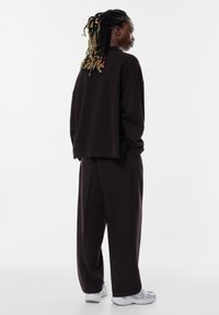 Oversized zwarte sweatshirt en bijpassende losse broek, beiden met een gestructureerde afwerking; gecombineerd met lichtgrijze sportschoenen.