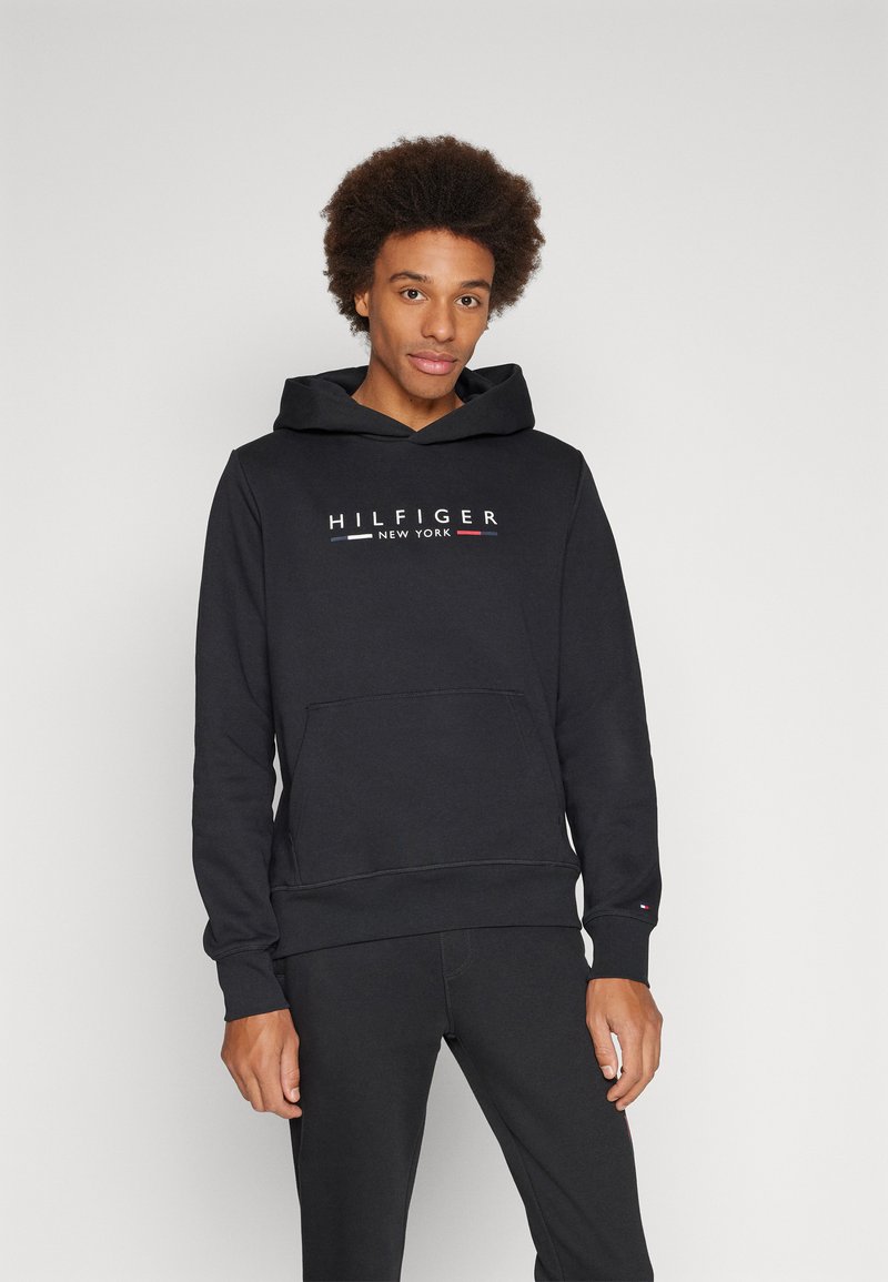 Crna dukserica od pamuka sa dodatkom, koja ima prednji džep i bijeli logo "HILFIGER NEW YORK" s crvenim i plavim akcentima.