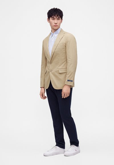Ung mand iført en beige blazer, blåstribet skjorte, mørke bukser og hvide sneakers står mod en ensfarvet lys baggrund.