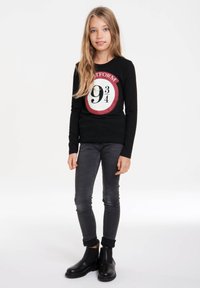 Schwarzes Langarmshirt mit einem runden roten und weißen Aufdruck mit "Plattform 9¾". Getragen mit grauen Skinny-Jeans und schwarzen Stiefeletten.