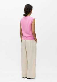 Mujer con cabello oscuro recogido en un moño bajo, lleva una camiseta sin mangas rosa y pantalones anchos beige, de pie con las manos en los bolsillos y mirando hacia atrás.
