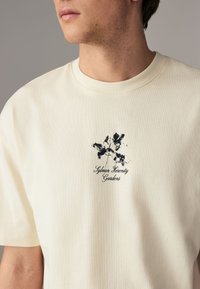 Cream cotton t-shirt featuring a black floral graphic with "Sylvan Serenity Gardens" text. Classic crew neck and short sleeves.