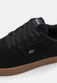 Black suede skate shoe featuring a textured upper, flat laces, and a rubber outsole with a natural brown finish. Logo on the tongue.