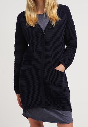 Frau trägt einen marineblauen, gerippten, langen Cardigan mit Fronttaschen über einem grauen Kleid, Hände in den Taschen, vor schlichtem Hintergrund.
