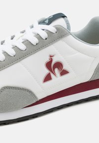 Baskets blanches avec des accents en daim gris, arborant un logo rouge, des lacets blancs et une semelle texturée. Des lignes de design subtiles sont visibles sur le matériau.