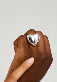 Sølv hjerteformet ring båret på en mørk hånd. Ringen har en glat, reflekterende overflade med et afrundet design og poleret finish.