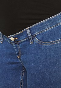 Denimové jeansy v tmavě modré barvě mají klasický design s pěti kapsami, měděné prošívání a kovové zavírání na pásu.