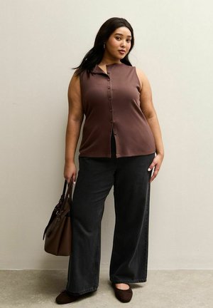 Top marrone senza maniche con chiusura a bottoni, abbinato a jeans neri a gamba larga. La modella tiene in mano una borsa marrone e indossa ballerine marrone scuro.