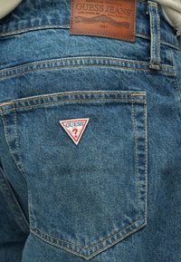 Niebieskie dżinsy z denimu z trójkątną czerwoną i białą naszywką z logo "GUESS" na tylnej kieszeni, ozdobione złotym przeszyciem i skórzaną metką na pasku w talii.