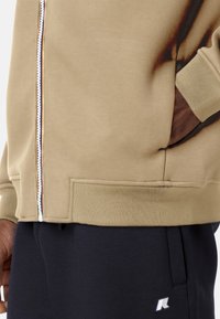 Giacca beige con zip, dettagli scuri, bordo e polsini a coste, tasche laterali e una texture liscia, indossata sopra pantaloni scuri con un logo.