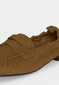 Mocassin en daim marron avec un arrière élastiqué, une bride décorative sur le dessus et des détails cousus sur les côtés.