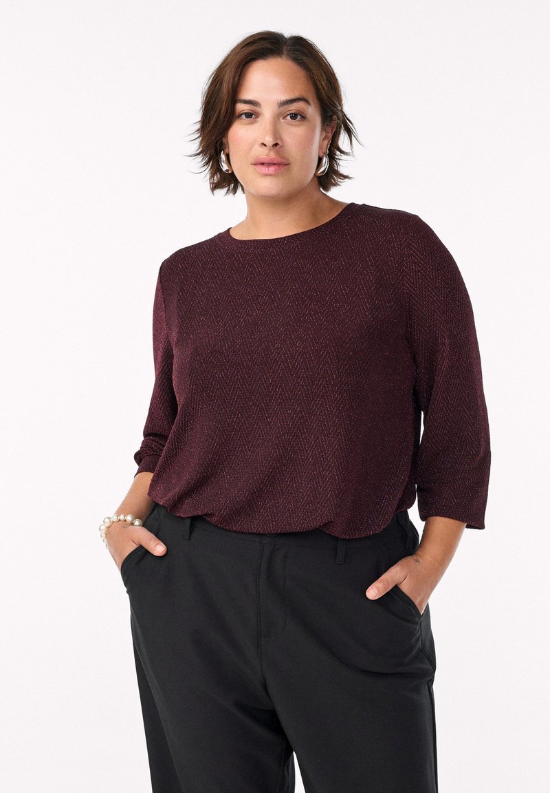 Bordová, textúrovaná blúzka s rybím vzorom, okrúhlym výstrihom a trištvrťovými rukávmi. Skombinovaná s čiernymi nohavicami a perlovým náramkom.