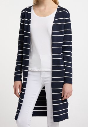 Personne portant un long cardigan bleu marine avec des rayures horizontales blanches sur une chemise blanche et un pantalon blanc, debout devant un fond blanc.