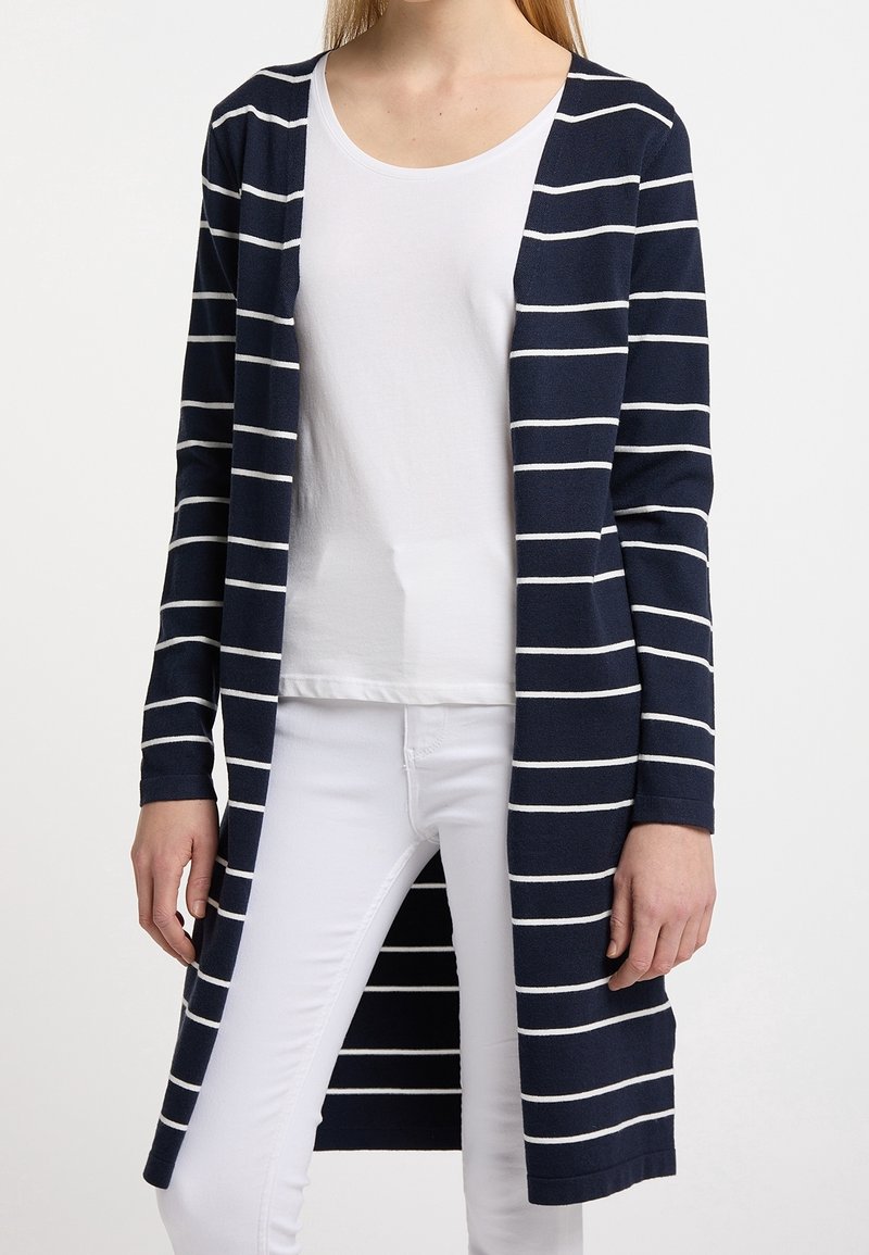 Personne portant un long cardigan bleu marine avec des rayures horizontales blanches sur une chemise blanche et un pantalon blanc, debout devant un fond blanc.
