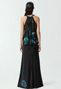 Crna cvjetna maksi haljina s prozirnim halter izrezom, koja ima mašnu oko vrata. Tkanina prikazuje zelene i ljubičaste uzorke cvijeća.