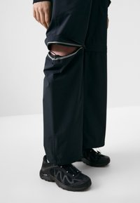 Zwarte waterdichte broek met ritssluitingen bij de knieën, gecombineerd met zwarte sportieve schoenen met gaasdetails en tekstuurlzolen.