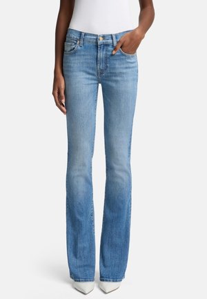 Femme portant un jean bootcut bleu clair et des chaussures pointues blanches, une main dans la poche et un haut blanc visible à la taille.