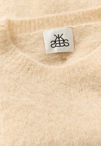 Cremfarbener Pullover mit einer flauschigen Textur und geripptem Ausschnitt. Verfügt über ein Etikett mit einem schwarzen Logo auf weißem Stoff.