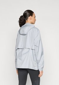 Veste coupe-vent gris clair avec capuche, au fit décontracté et aux poignets élastiques, remarquable par son design minimaliste et son étiquette sur le côté.