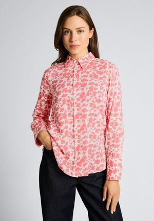 Vrouw met lang bruin haar, gekleed in een roze-wit bloes met knopen en een donkere broek, staand met één hand in haar zak.